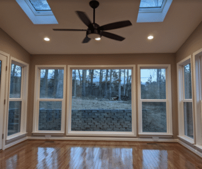 A sunroom with a dark wood floor, large windows overlooking a wooded area, a ceiling fan, and two skylights.