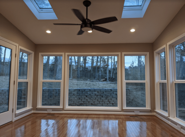 A sunroom with a dark wood floor, large windows overlooking a wooded area, a ceiling fan, and two skylights.